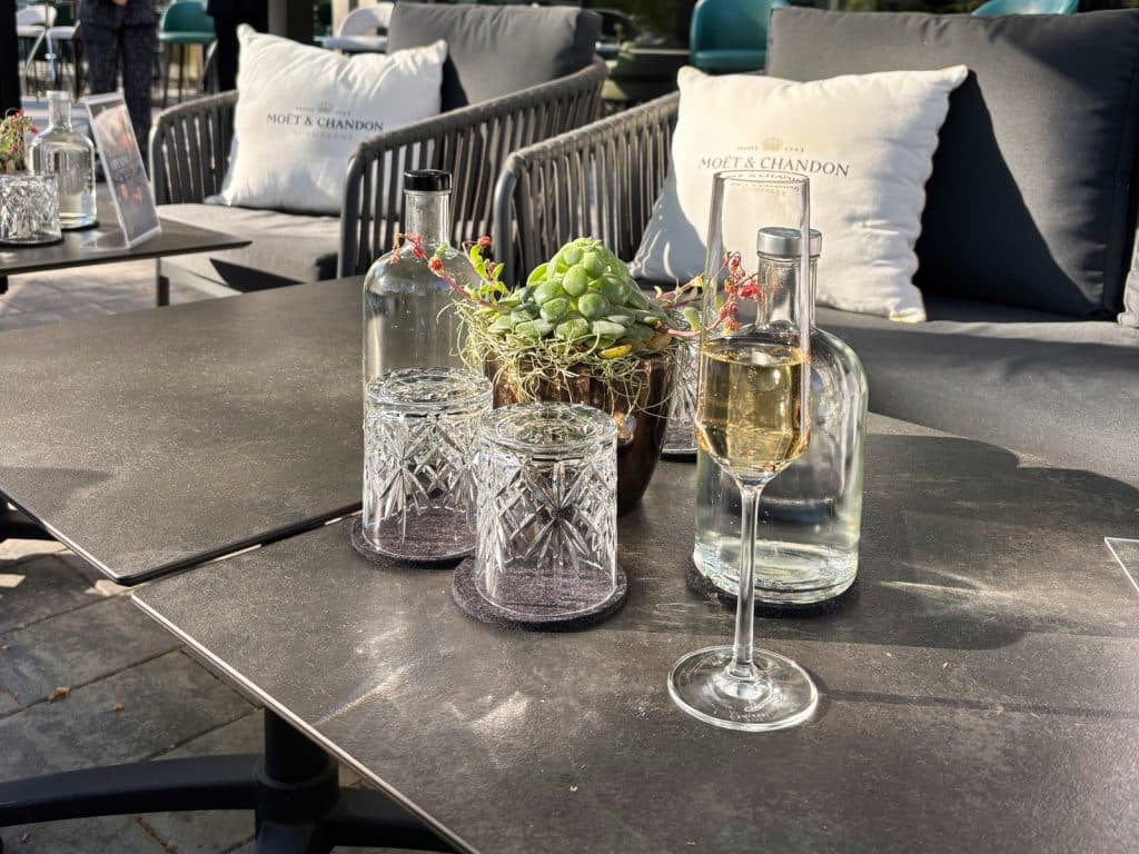Ein gedeckter Tisch auf der Terrasse des DoubleTree by Hilton Berlin Ku'damm mit einem Glas Champagner, Wasserflaschen und Kristallgläsern vor gemütlichen Loungesesseln mit Moët & Chandon Kissen.
