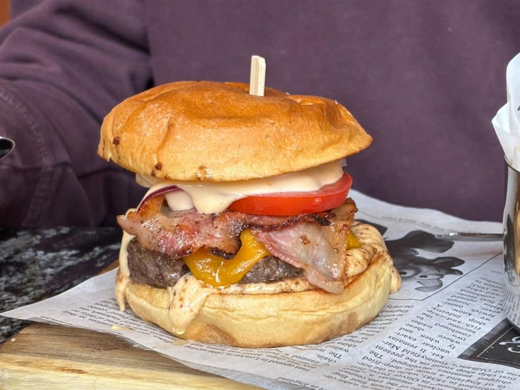 Ein saftiger BESADA-Burger im Restaurant BESADA in Pilsen, belegt mit Rindfleisch, geschmolzenem Käse, krossem Speck, Tomate und Sauce in einem Brioche-Brötchen, serviert auf bedrucktem Papier.