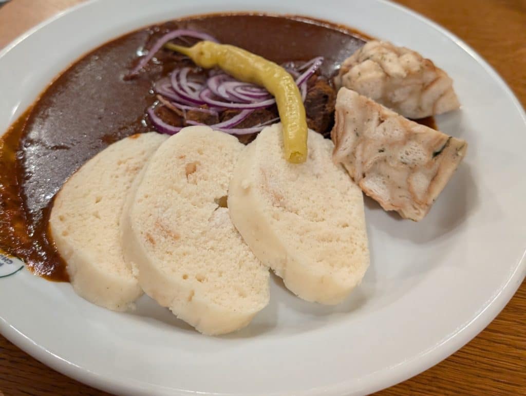 Ein Teller mit traditionellem tschechischem Gulasch im Restaurant Beseda in Pilsen, serviert mit hellen böhmischen Knödeln, Karlsbader Knödeln, roten Zwiebelringen und einer Peperoni.