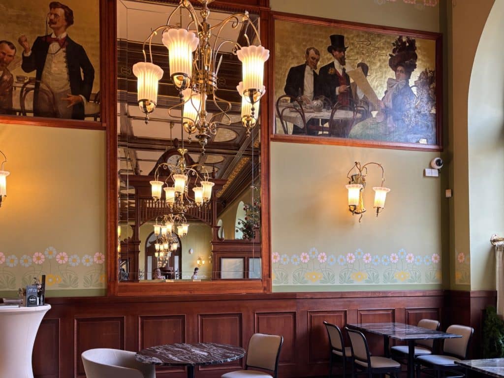 Innenansicht des traditionellen tschechischen Restaurants BESADA in Pilsen mit holzgetäfelten Wänden, kunstvollen Wandgemälden im Belle-Époque-Stil, einem großen Spiegel und eleganten Kronleuchtern über Tischen mit Marmorplatte.