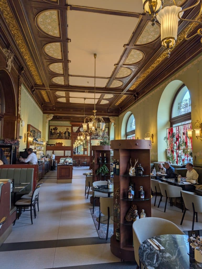 Der prachtvolle Innenraum des traditionellen tschechischen Restaurants Beseda in Pilsen besticht durch eine aufwendig verzierte Holzkassettendecke, elegante Kronleuchter und gemütliche Sitzbereiche an runden Tischen. Große Rundbogenfenster lassen Tageslicht in den hohen Gastraum.