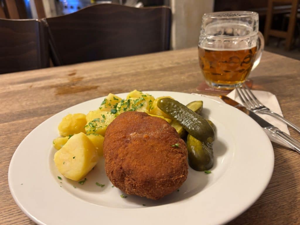 Ein Teller mit traditionellem tschechischem Essen in einem Restaurant in Pilsen, bestehend aus einem panierten Fleischgericht, Petersilienkartoffeln und Gewürzgurken, serviert neben einem Glas Pilsener Bier.