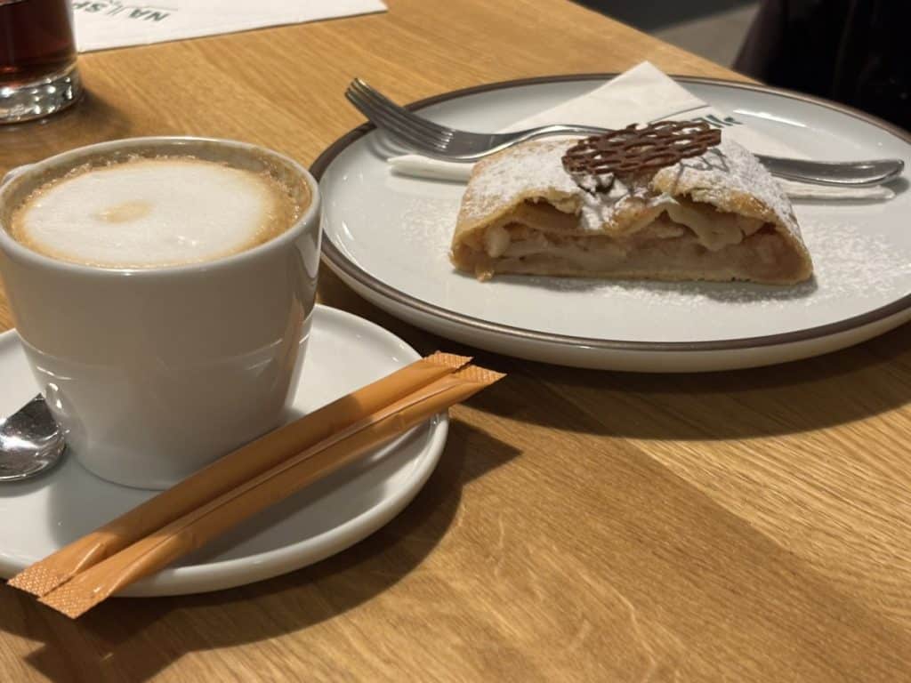 Ein Stück traditioneller tschechischer Apfelstrudel mit Puderzucker und Schokoladendekor auf einem Teller neben einer Tasse Kaffee im Restaurant Na Spilce in Pilsen.