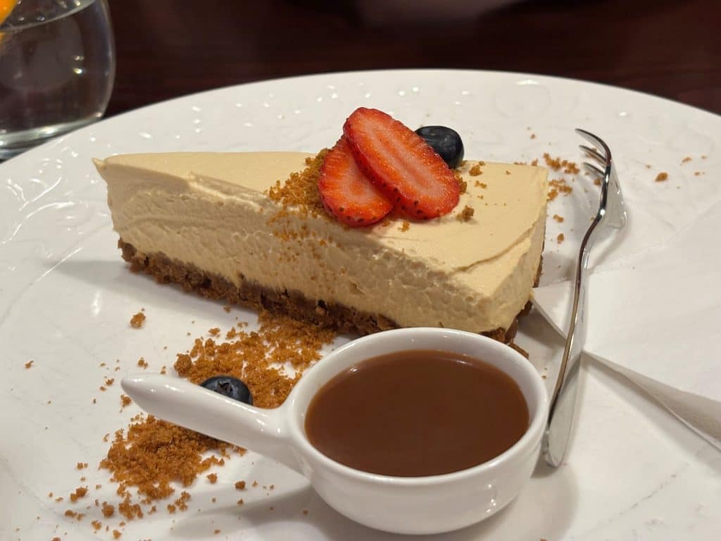 Ein Stück traditioneller tschechischer Kuchen im Restaurant Purkmistr in Pilsen, garniert mit Erdbeerscheiben, Blaubeeren und Keksbröseln, serviert mit einer kleinen Schale Karamellsauce auf einem weißen Teller.