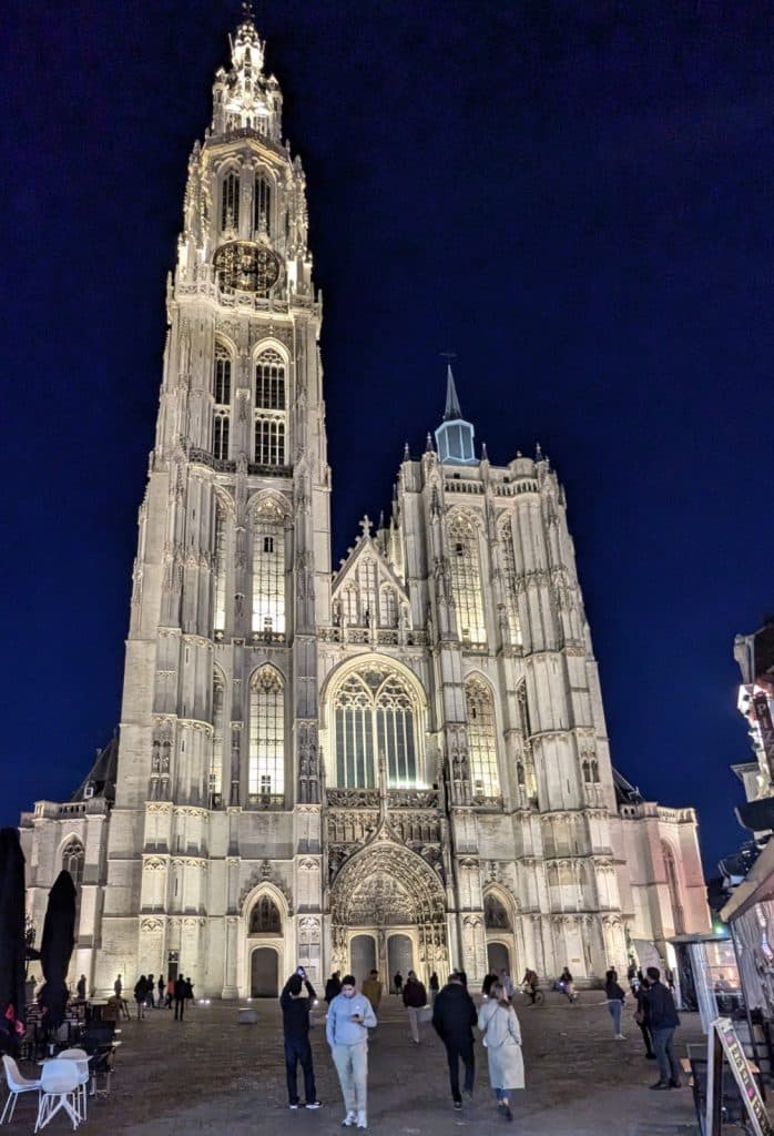 Die beleuchtete Fassade der Kathedrale Unserer Lieben Frau in Antwerpen ragt bei Nacht hinter einem belebten Platz mit Passanten in den dunklen Himmel.