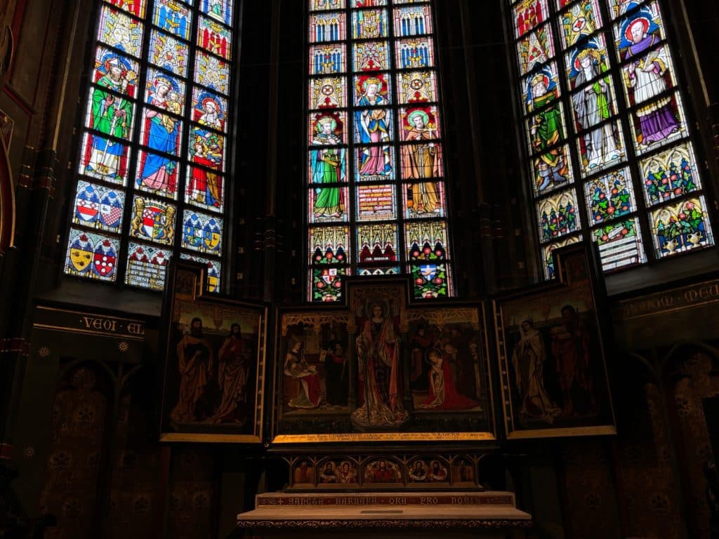 Ein prachtvoller Altar in der Liebfrauenkathedrale von Antwerpen, über dem drei hohe, farbenfrohe Buntglasfenster mit religiösen Figuren und Wappen thronen. Das zentrale Triptychon zeigt detaillierte sakrale Malereien in einem dunklen, stimmungsvollen Kirchenraum.