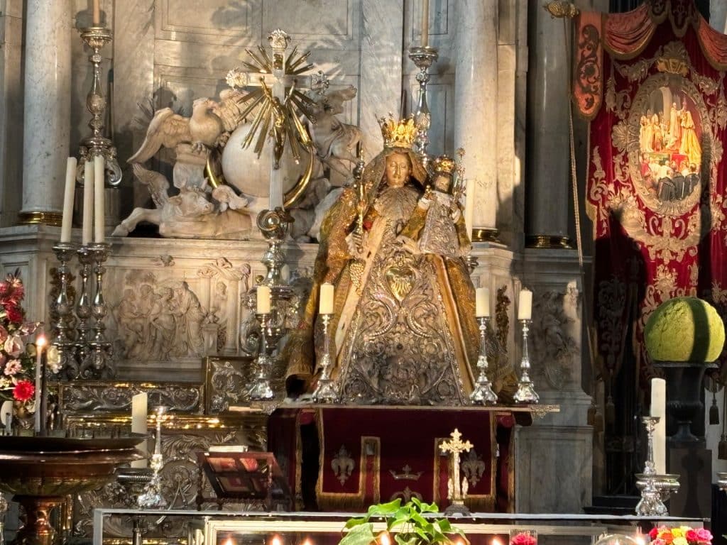 Eine prunkvoll gekleidete Madonna mit Kind steht auf einem silbernen Altar in der Kathedrale von Antwerpen, umgeben von Kerzenleuchtern, religiösen Skulpturen und einem roten Banner.