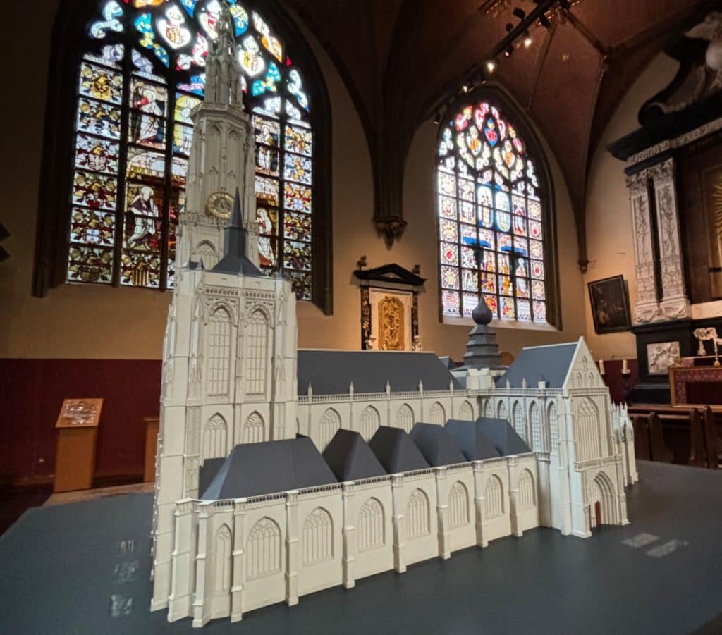 Ein detailliertes, weißes Architekturmodell der Antwerpener Liebfrauenkathedrale steht in einem Kirchenraum vor farbenprächtigen Buntglasfenstern. Das Modell zeigt die gotische Struktur mit dem markanten hohen Turm, dem Kirchenschiff und den filigranen Fassadendetails.