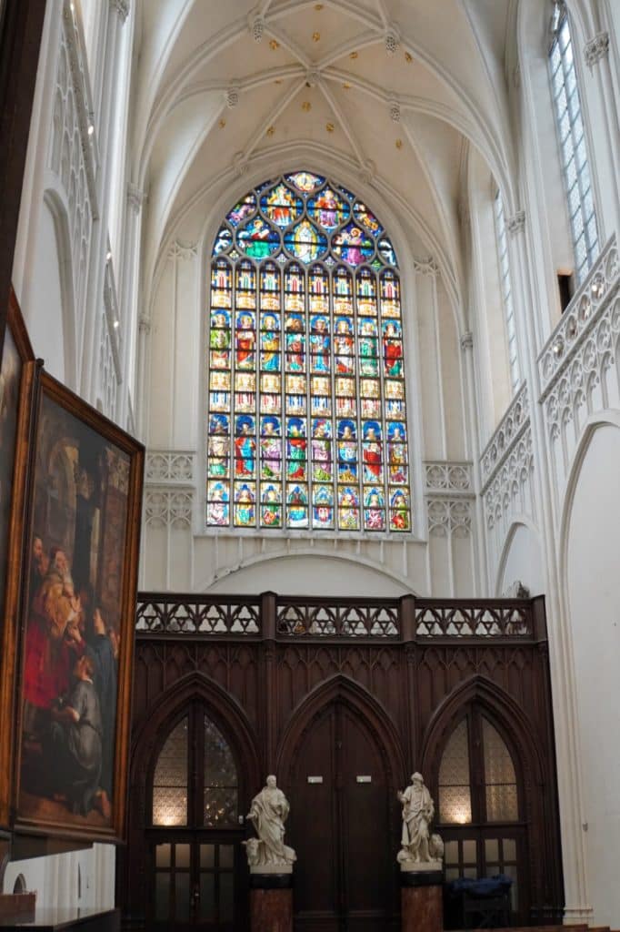 Blick in das helle Querschiff der Kathedrale von Antwerpen mit einem großen, farbenfrohen Buntglasfenster über einer dunklen Holzempore mit Statuen und einem Gemälde im Vordergrund.