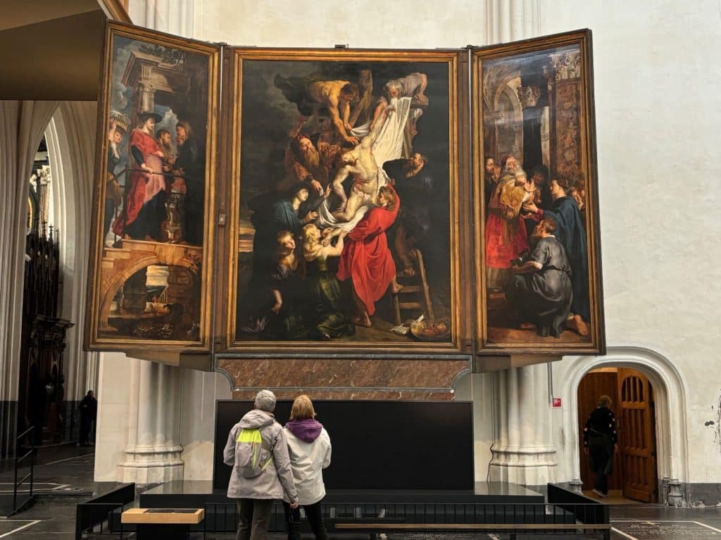 Zwei Personen betrachten das monumentale Triptychon „Die Kreuzabnahme“ von Peter Paul Rubens, das in einem prunkvollen Goldrahmen über einem Altar präsentiert wird.