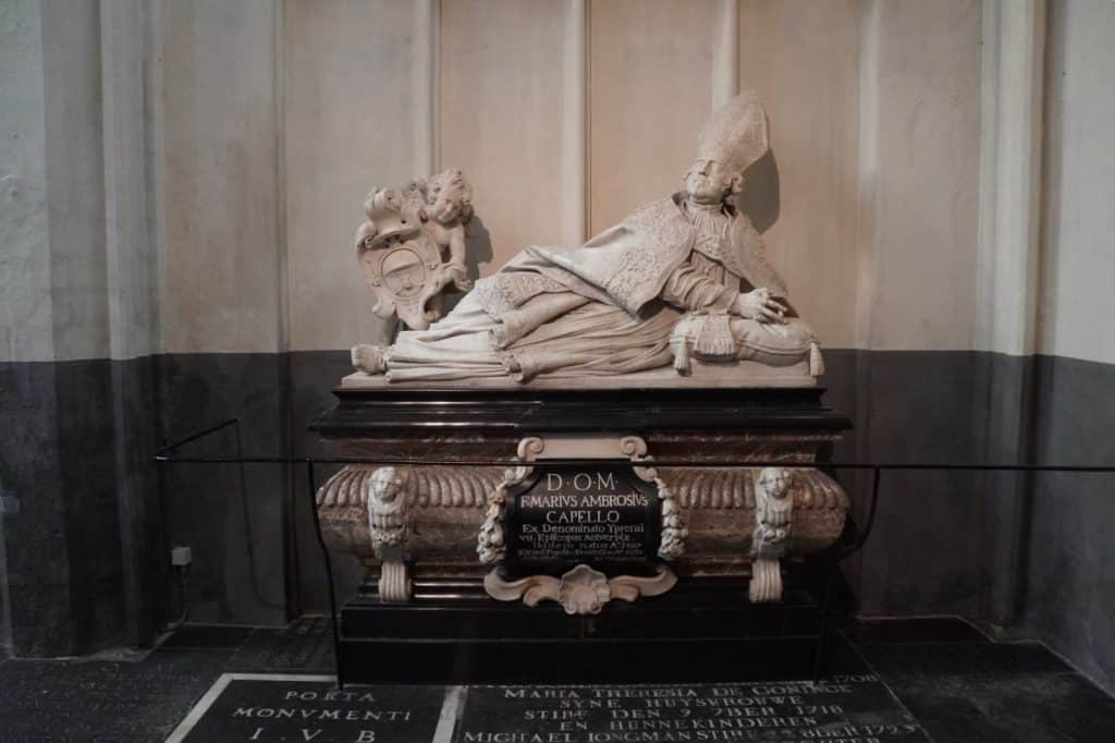 Ein prunkvoller barocker Sarkophag in der Kathedrale von Antwerpen, auf dem eine weiße Marmorskulptur des Bischofs Marius Ambrosius Capello in liegender, aufgestützter Pose ruht. Das Grabmal besteht aus einem dunklen Sockel mit einer lateinischen Inschriftentafel und wird von einem kleinen Engel flankiert, der ein Wappen hält.