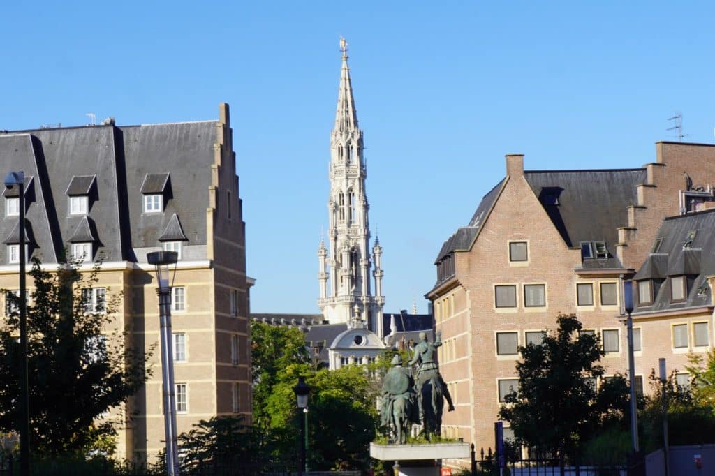 Blick durch eine Häuserzeile auf den filigranen Turm der Kathedrale von Antwerpen unter strahlend blauem Himmel, im Vordergrund steht ein Reiterstandbild.