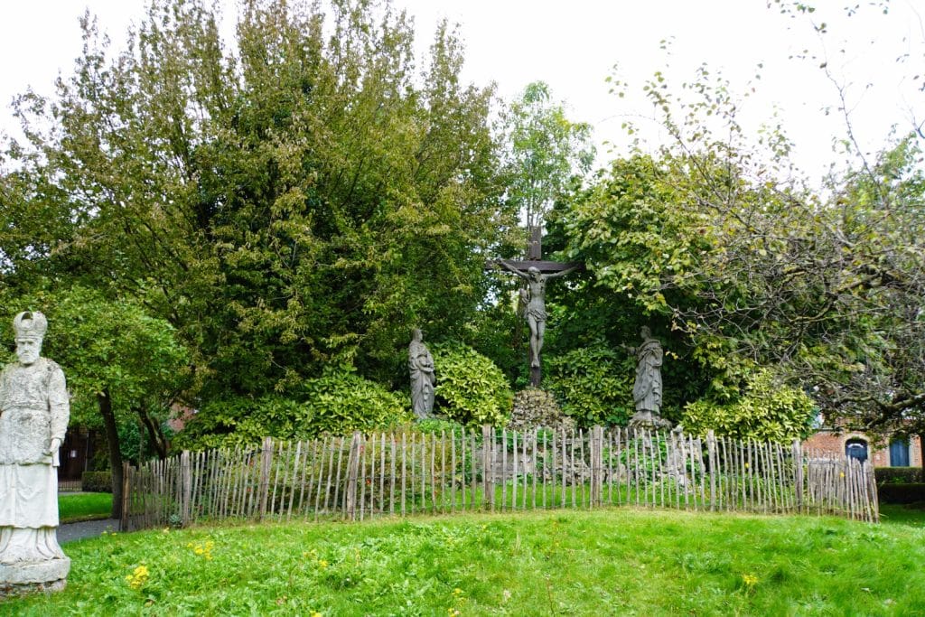 Das Bild zeigt eine religiöse Stätte im Beginenhof in Antwerpen, umgeben von üppigem Grün. Im Zentrum steht ein Kreuz mit einer Christusfigur, flankiert von Statuen und einem niedrigen Holzzaun. Eine weitere Statue, möglicherweise eines Heiligen, steht links im Bild.