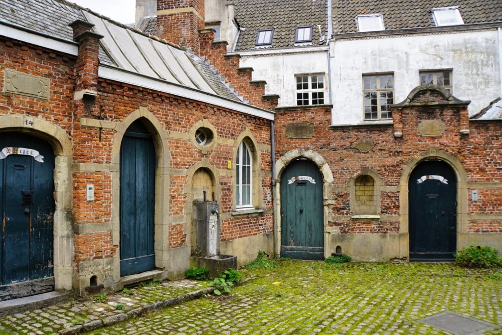 Das Bild zeigt den Beginenhof in Antwerpen mit mehreren Hauseingängen aus rotem Backstein, die sich um einen Innenhof mit Kopfsteinpflaster gruppieren. Die Türen sind in verschiedenen Grüntönen gehalten und mit weißen Bändern über den Eingängen verziert.