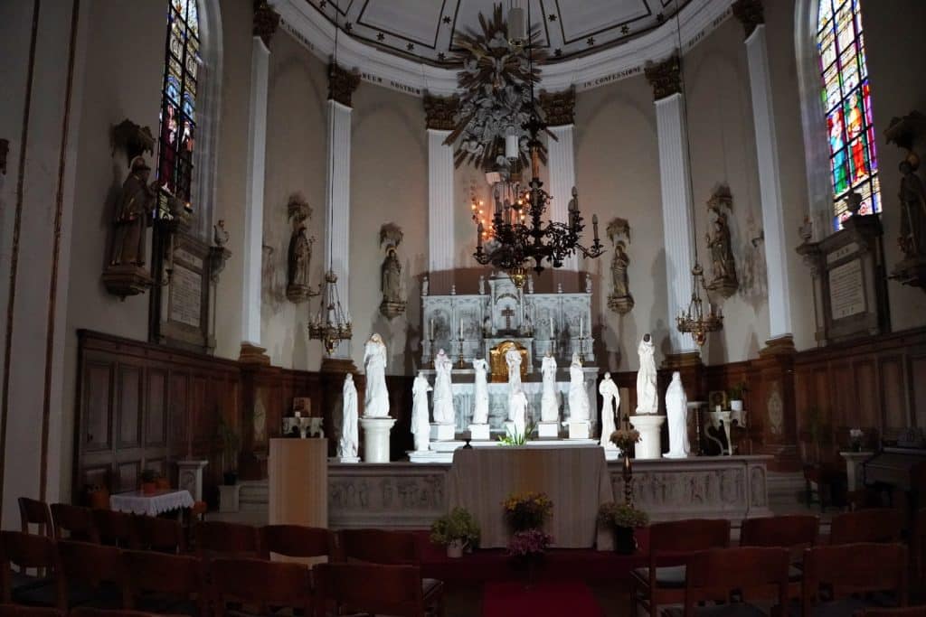 Innenansicht der Kirche Beginenhof in Antwerpen mit Blick auf den Altarraum, der mit weißen Statuen geschmückt ist, und den Kirchenraum mit hölzernen Kirchenbänken.