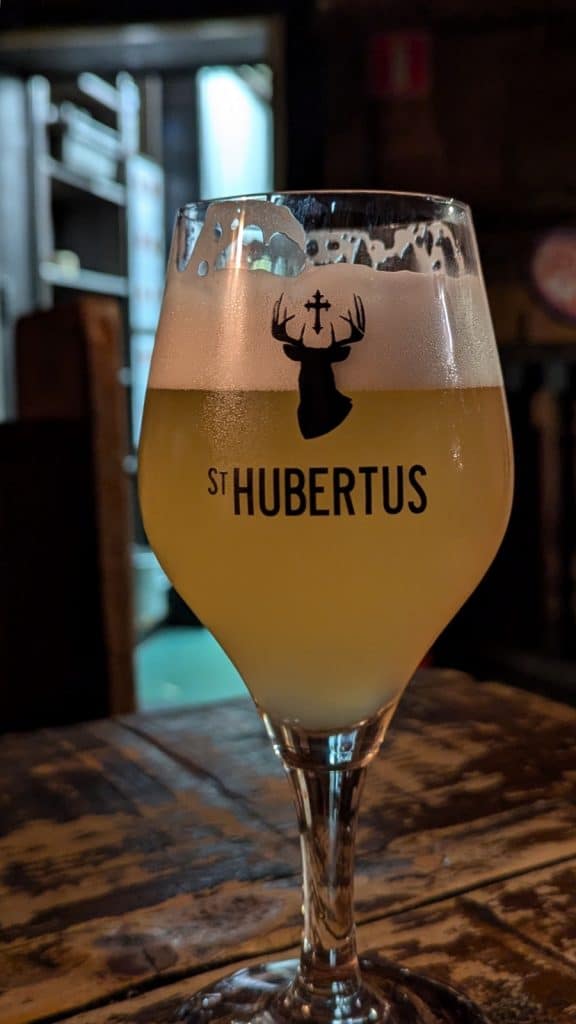 Ein Glas helles, naturtrübes St. Hubertus Bier mit weißer Schaumkrone steht auf einem rustikalen Holztisch im Bier Central in Antwerpen. Das Glas zeigt das schwarze Logo eines Hirschkopfs mit einem Kreuz zwischen dem Geweih und den Schriftzug „ST HUBERTUS“.