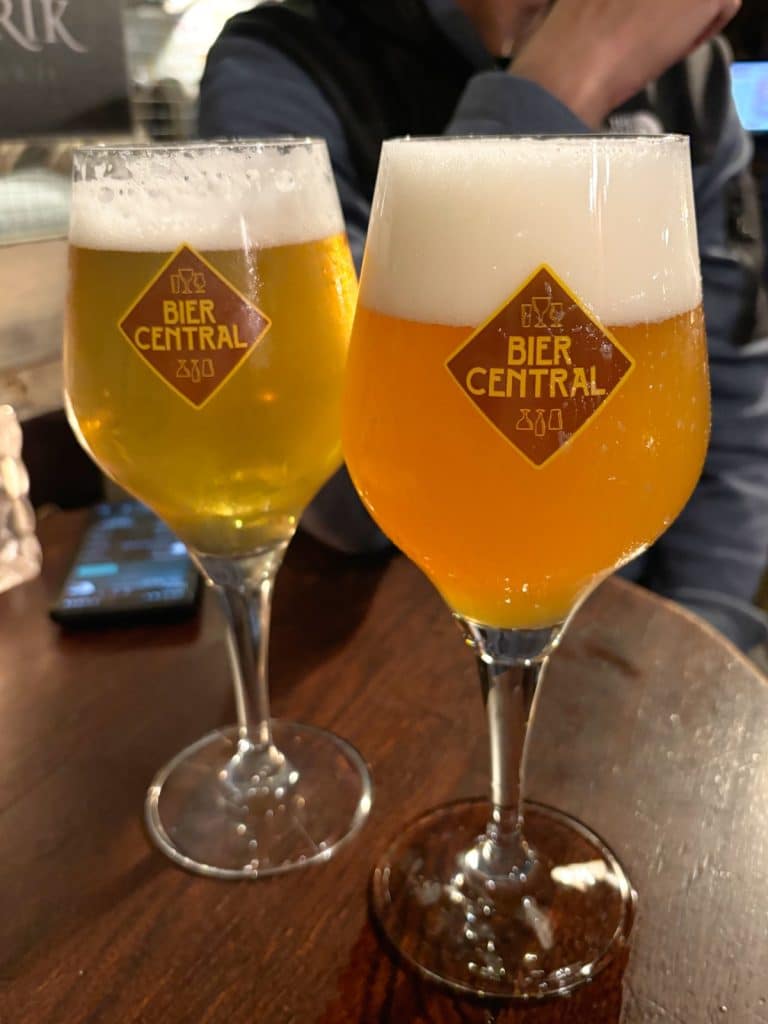 Zwei Gläser mit hellem und trübem Bier der Bier Central in Antwerpen stehen auf einem dunklen Holztisch, wobei das Logo der Bar deutlich auf den Kelchgläsern zu sehen ist.