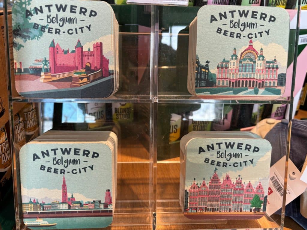 Vier Stapel quadratischer Bierdeckel mit der Aufschrift „Antwerp - Belgium - Beer-City“ zeigen in einem Regal verschiedene illustrierte Wahrzeichen der Stadt Antwerpen in Rosa- und Blautönen. Jedes Motiv präsentiert eine andere Sehenswürdigkeit, darunter die Burg Het Steen, den Hauptbahnhof, die Skyline mit Kathedrale und die historischen Gildehäuser am Grote Markt.