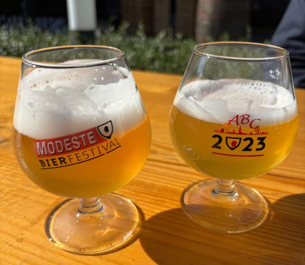 Zwei Gläser helles Bier mit Schaumkrone stehen auf einem Holztisch beim Modeste Bierfestival in Antwerpen, wobei die Gläser Logos des Festivals tragen.