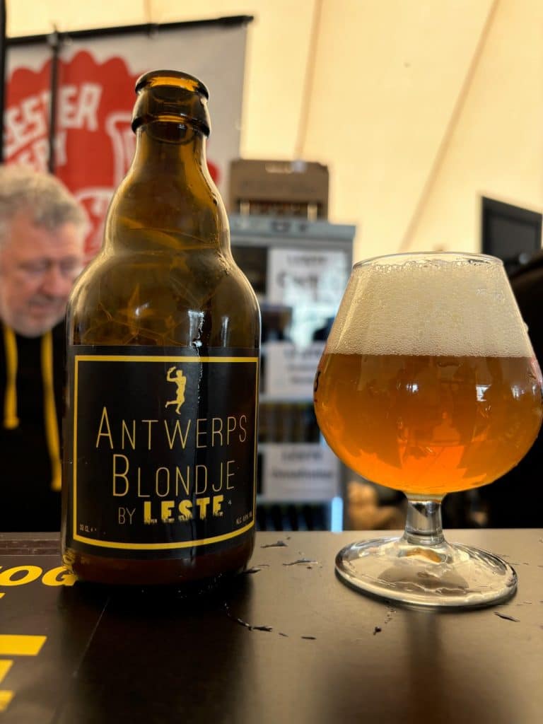Eine braune Bierflasche mit dem Etikett „Antwerps Blondje by Leste“ steht neben einem gefüllten Glas mit hellem Bier und weißer Schaumkrone auf dem Antwerpener Bierfestival.