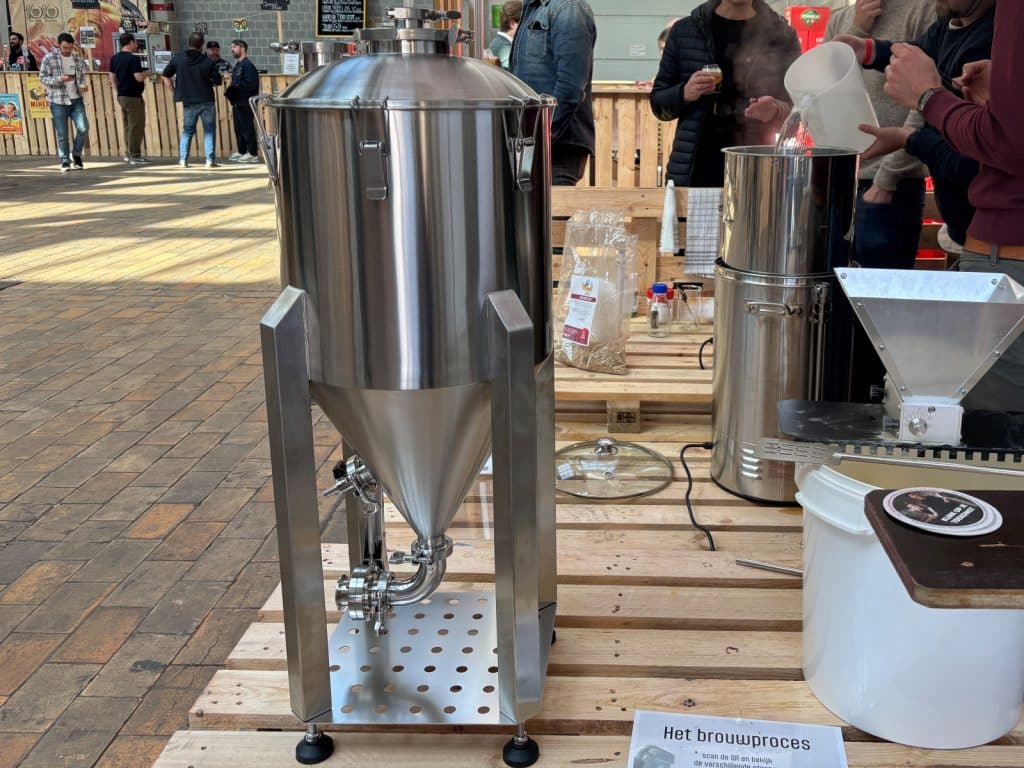 Ein glänzendes Brauset aus Edelstahl mit Gärtank und Malzmühle steht auf Holzpaletten während des Antwerpen Bier Festivals, während im Hintergrund Besucher das Brauverfahren beobachten.