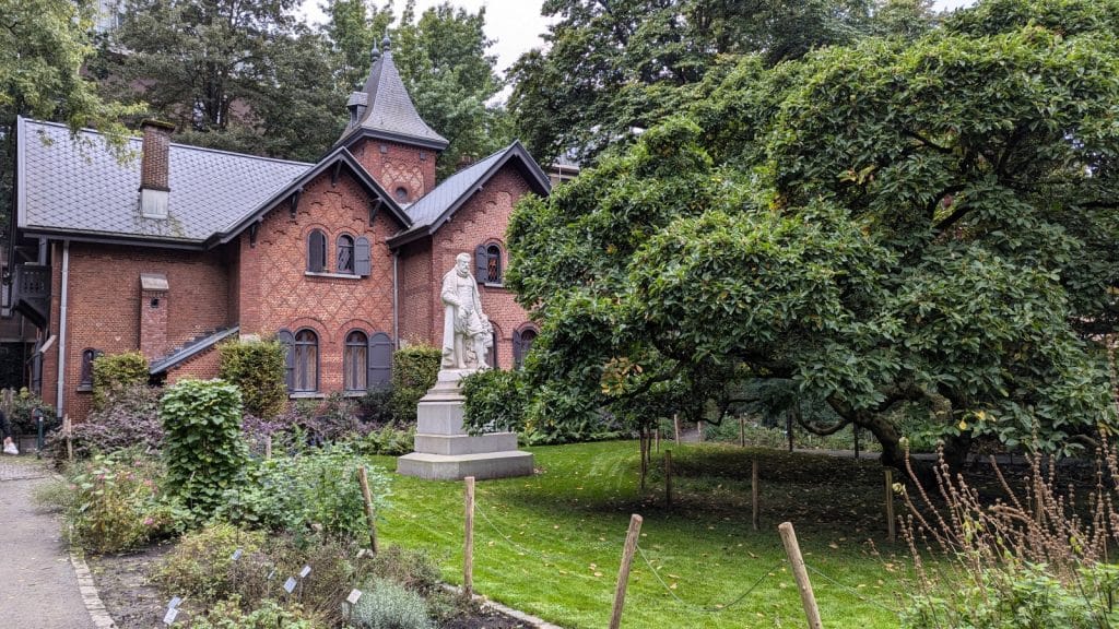 Das Bild zeigt ein Gebäude im Botanischen Garten von Antwerpen, das sich durch seine rote Backsteinfassade und den kleinen Turm auszeichnet. Vor dem Gebäude befindet sich eine Statue auf einem Sockel, umgeben von gepflegtem Rasen und üppigen Bäumen.