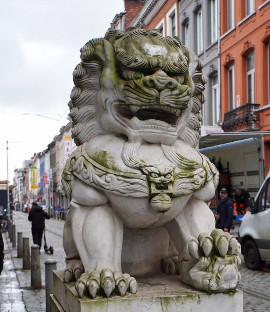 Eine massive, wettergegerbte Steinskulptur eines chinesischen Drachenlöwen (Wächterlöwe) steht auf einem Sockel in einer belebten Straße im Chinatown-Viertel von Antwerpen. Im Hintergrund sind Passanten, ein weißer Lieferwagen und die charakteristischen Fassaden europäischer Stadthäuser unter einem bewölkten Himmel zu sehen.