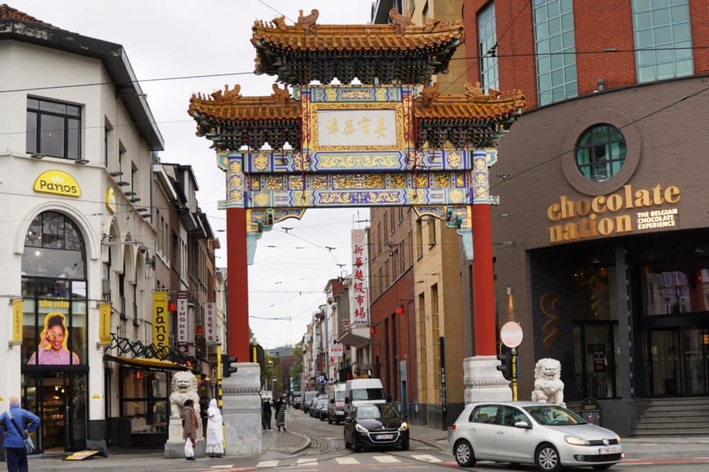 Ein großes, farbenfrohes chinesisches Tor mit goldenen Verzierungen und Pagodendach überspannt den Eingang zum Viertel Chinatown in Antwerpen, flankiert von modernen Gebäuden wie dem Museum „Chocolate Nation“. Vor dem Tor stehen zwei traditionelle Löwenstatuen aus Stein, während Autos und Fußgänger die belebte Straße passieren.