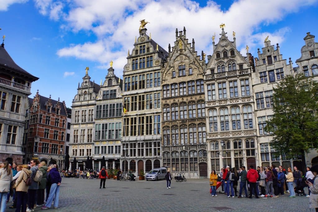 Das Bild zeigt den Grote Markt in Antwerpen mit den historischen Zunfthäusern, die sich durch ihre reich verzierten Fassaden und goldenen Statuen auszeichnen. Im Vordergrund sind Gruppen von Menschen zu sehen, die den Platz bevölkern, während der Himmel mit weißen Wolken bedeckt ist.