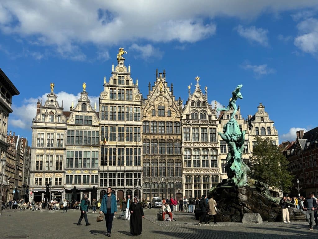 Das Bild zeigt den Grote Markt in Antwerpen, einen belebten Platz mit historischen Gildehäusern und dem Brabo-Brunnen im Vordergrund. Menschen flanieren über den Platz, einige sitzen in den Außenbereichen der Cafés und Restaurants.
