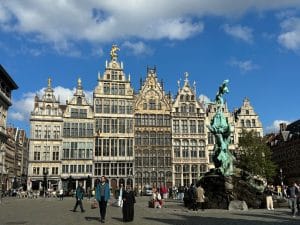 Das Bild zeigt den Grote Markt in Antwerpen, einen belebten Platz mit historischen Gildehäusern und dem Brabo-Brunnen im Vordergrund. Menschen flanieren über den Platz, einige sitzen in den Außenbereichen der Cafés und Restaurants.