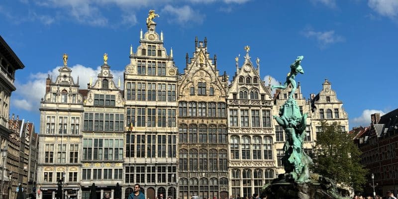 Das Bild zeigt den Grote Markt in Antwerpen, einen belebten Platz mit historischen Gildehäusern und dem Brabo-Brunnen im Vordergrund. Menschen flanieren über den Platz, einige sitzen in den Außenbereichen der Cafés und Restaurants.