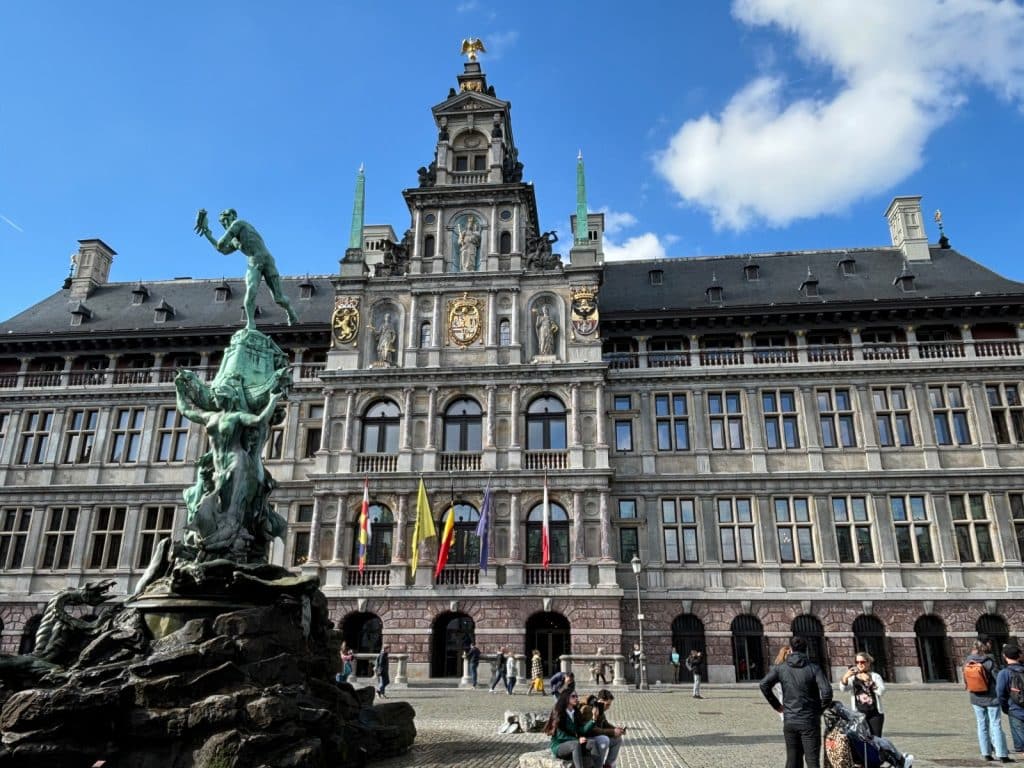 Das Bild zeigt das Rathaus von Antwerpen auf dem Grote Markt, mit einer Statue im Vordergrund und Menschen, die sich auf dem Platz aufhalten. Das Rathaus ist ein großes, verziertes Gebäude mit mehreren Etagen und einem Turm.