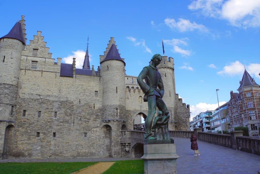 Vor dem mittelalterlichen Eingang der Burg Het Steen in Antwerpen steht eine große Bronzestatue des Riesen Lange Wapper auf einer Brücke. Die historische Festung aus hellem Stein zeigt markante Türme mit spitzen Dächern unter einem blauen Himmel mit lockeren Wolken.