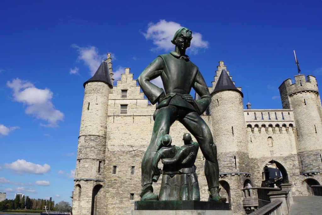 Das Bild zeigt die Statue von Lange Wapper vor dem Het Steen in Antwerpen. Die Statue stellt einen Riesen dar, der über zwei kleinere Figuren steht, während im Hintergrund das mittelalterliche Schloss Het Steen mit seinen Türmen und Zinnen zu sehen ist.