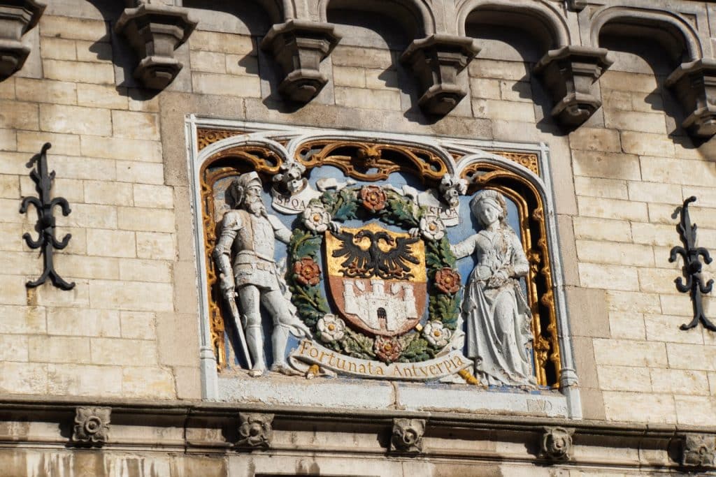 Ein farbenfrohes Relief an der Fassade der Burg Het Steen in Antwerpen zeigt das Stadtwappen mit einem doppelköpfigen Adler und einer Burg, flankiert von einer Ritterfigur und einer Frauengestalt unter dem Schriftzug „Fortunata Antverpia“.