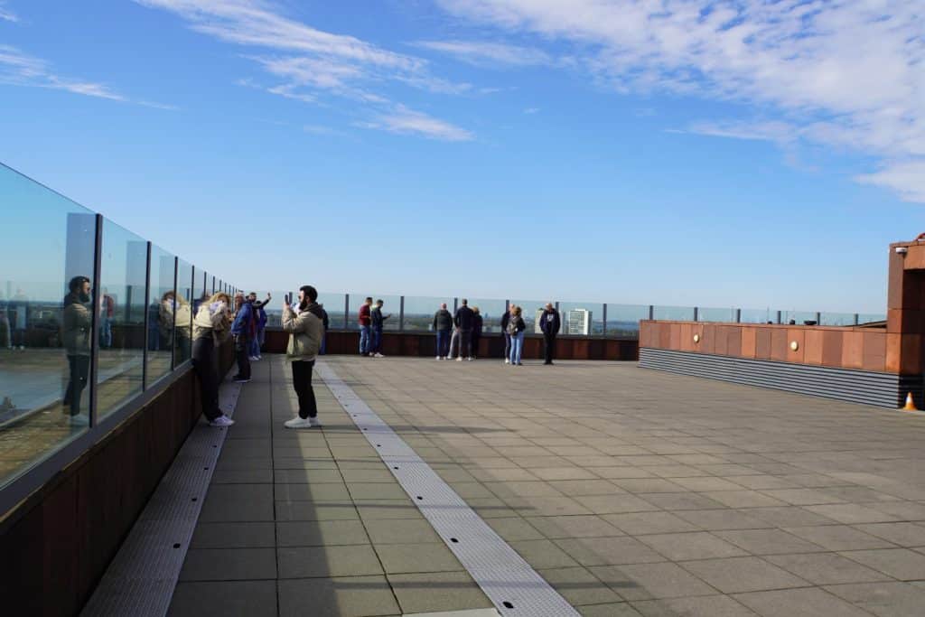 Besucher stehen auf der weitläufigen, gepflasterten Aussichtsplattform des MAS in Antwerpen hinter einer Glasbrüstung und genießen bei blauem Himmel den Blick über die Stadt.