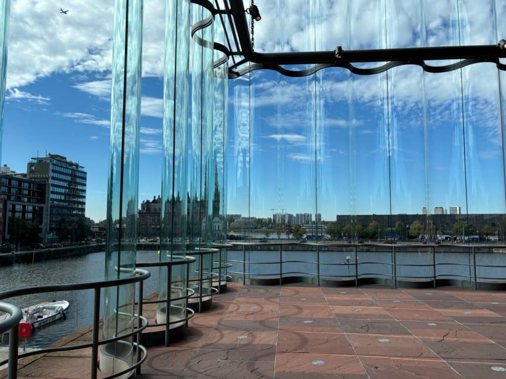 Blick aus dem Inneren des MAS in Antwerpen durch eine gewellte Glasfassade auf den Hafen, ein kleines Boot und die umliegende Stadtarchitektur unter blauem Himmel.