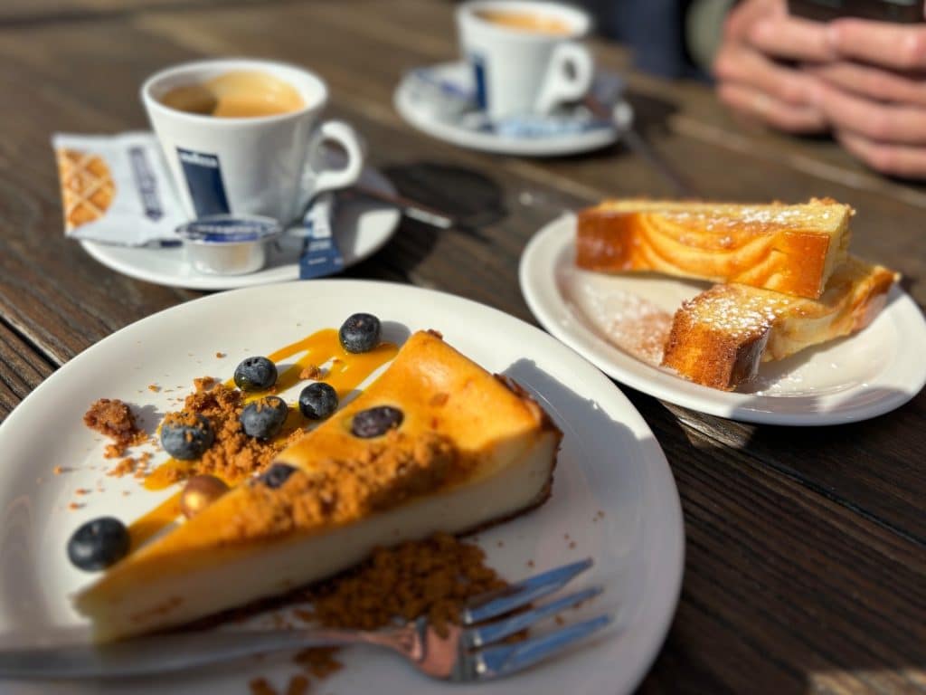 Ein Stück Käsekuchen mit Blaubeeren und Streuseln sowie zwei Scheiben Marmorkuchen stehen neben zwei Tassen Kaffee auf einem Holztisch im Freien beim MAS in Antwerpen.