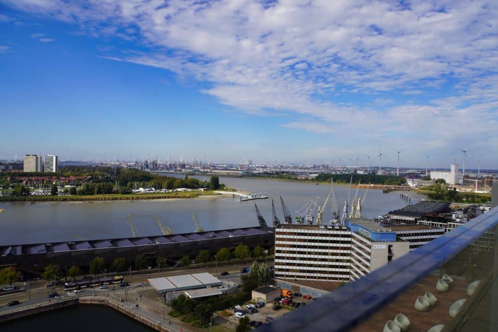 Blick vom Museum aan de Stroom (MAS) in Antwerpen über die Schelde auf den Hafen mit seinen Kränen, Industrieanlagen und Windrädern unter einem leicht bewölkten Himmel.