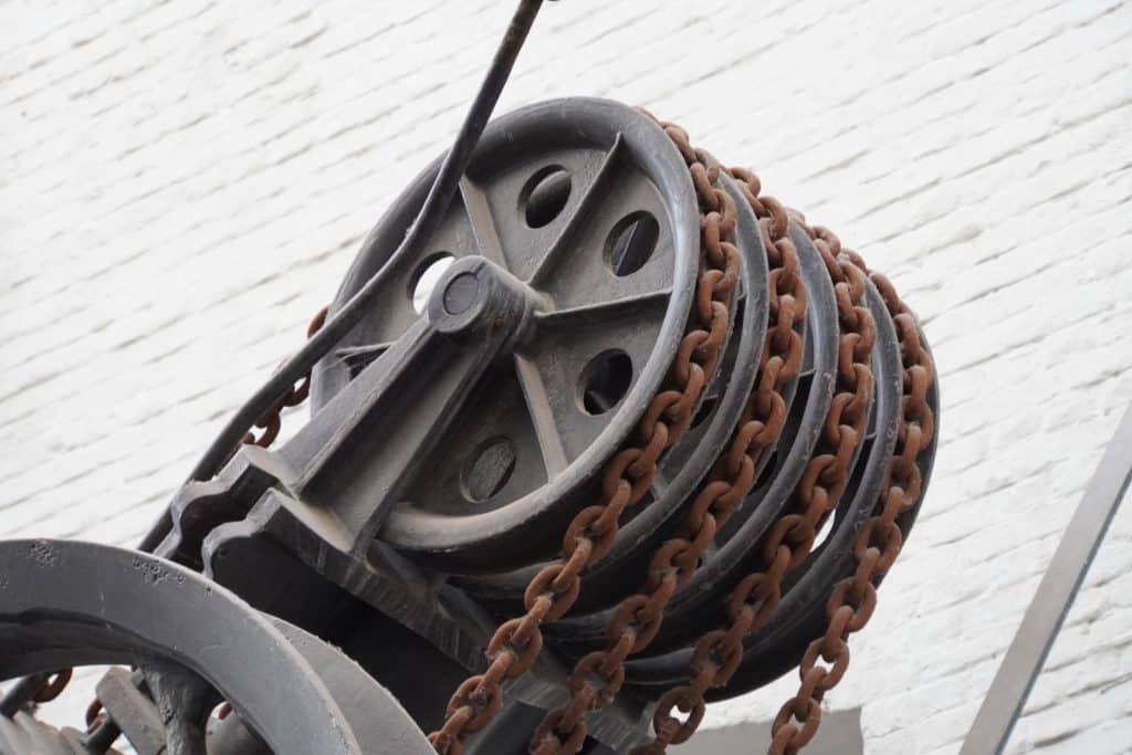 Ein historisches Detail in einer Passage in Antwerpen zeigt eine Nahaufnahme eines gusseisernen Flaschenzugs mit mehreren rostigen Ketten, die über schwere Metallrollen laufen.
