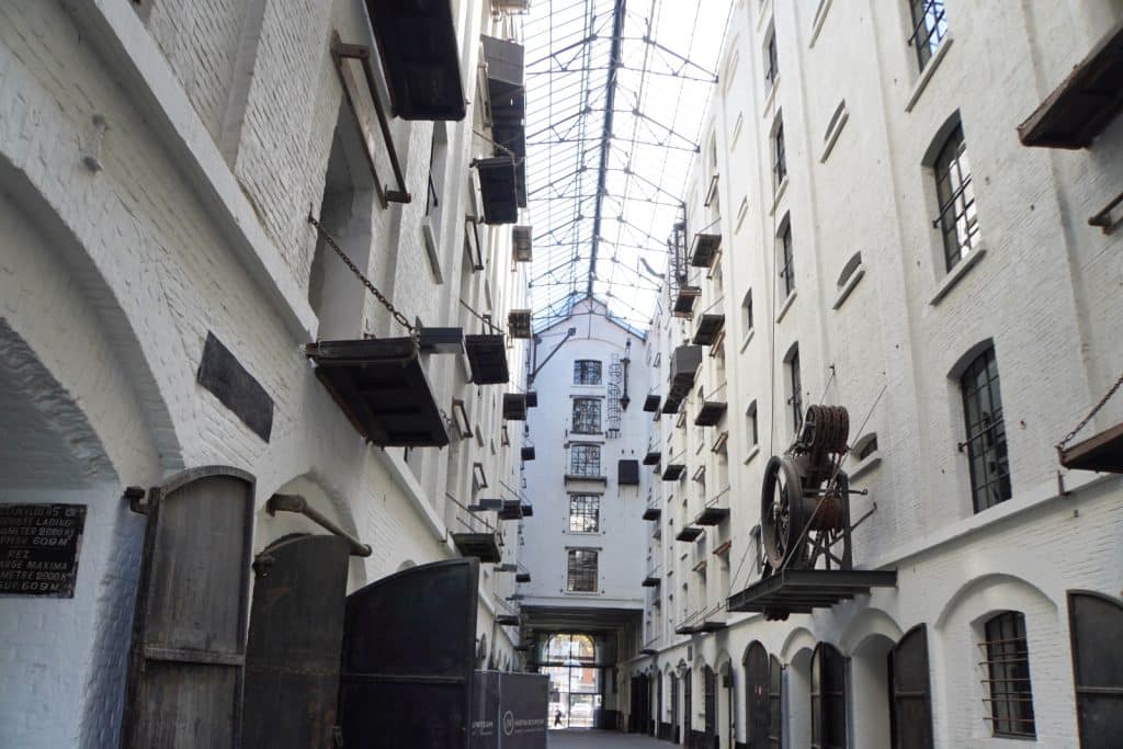 Blick durch die schmale, von weißen Backsteinfassaden gesäumte Passage quer durch einen historischen Speicherkomplex in Antwerpen, die von einem hohen Glasdach überspannt wird und durch zahlreiche Ladeklappen sowie eine alte Seilwinde geprägt ist.