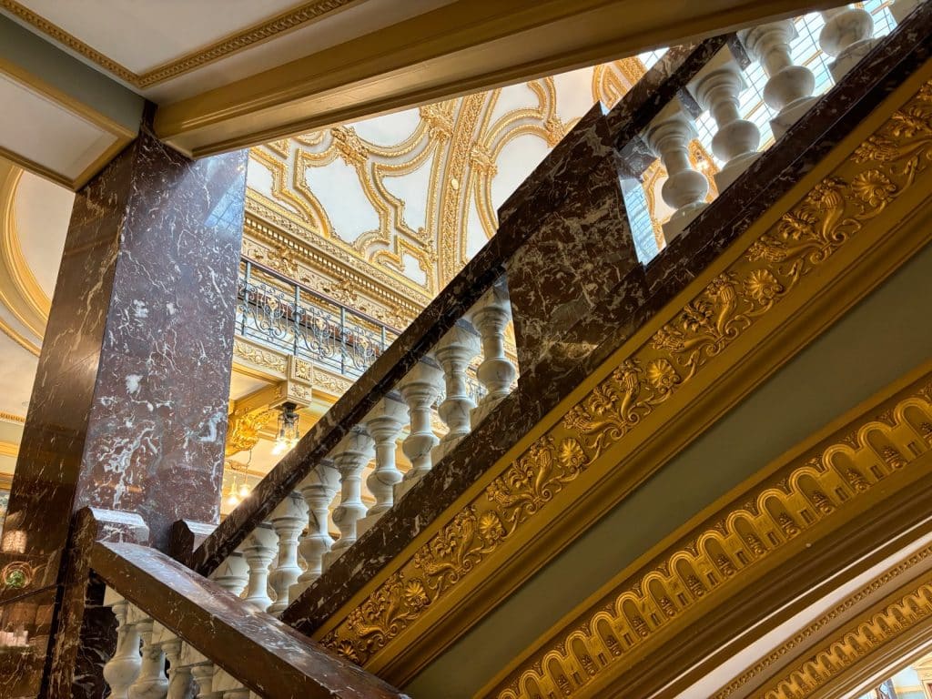 Dieses Foto zeigt das Treppengeländer der Stadsfestzaal in Antwerpen. Das Geländer besteht aus weißlichen Balustern und ist mit braunem Marmor und goldenen Verzierungen versehen.
