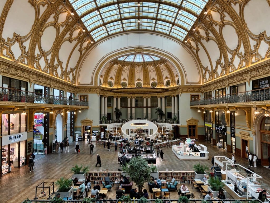 Das Innere der Stadsfeestzaal in Antwerpen zeigt eine weitläufige Halle mit Geschäften und Cafés, überdacht von einer beeindruckenden Kuppel mit goldenen Verzierungen und einem Glasdach, das Tageslicht hereinlässt. Menschen flanieren durch den Raum, sitzen an Tischen oder besuchen die Geschäfte.