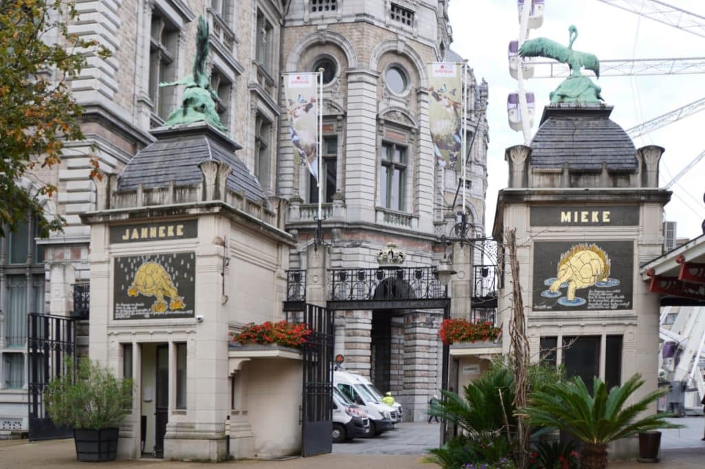 Das Bild zeigt den historischen Eingang zum Antwerpener Zoo, flankiert von zwei Torhäusern, die mit den Namen "Janneke" und "Mieke" beschriftet sind und jeweils ein Mosaik einer Schildkröte tragen. Über den Torhäusern befinden sich Skulpturen, und im Hintergrund ist ein Teil des Zoos mit einem Riesenrad zu sehen.
