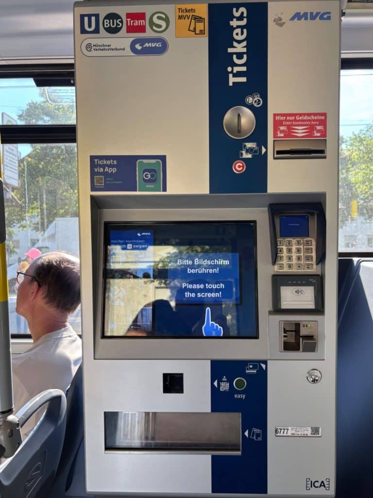 Ein moderner Fahrkartenautomat der MVG in einem Münchner öffentlichen Verkehrsmittel mit Touchscreen, Kartenzahlungsmodul und Münzeinwurf. Das Display zeigt die Aufforderung „Bitte Bildschirm berühren!“ auf Deutsch und Englisch an.