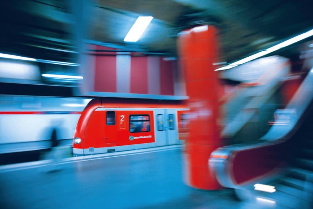 Eine dynamische Langzeitbelichtung einer einfahrenden S-Bahn in einer Münchener U-Bahn-Station, die durch Bewegungsunschärfe und leuchtend rote Akzente an Zug, Säulen und Rolltreppen geprägt ist.