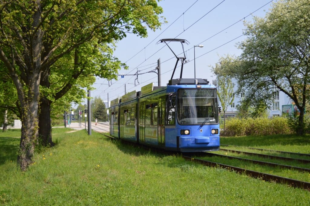 Eine blaue Münchener Straßenbahn fährt an einem sonnigen Tag auf Gleisen durch eine grüne Parklandschaft mit Bäumen.