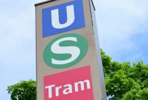 Ein Hinweisschild am Stachus in München zeigt untereinander die Symbole für die U-Bahn (weißes U auf blauem Grund), die S-Bahn (weißes S im grünen Kreis) und die Tram (weißer Schriftzug auf rotem Grund).