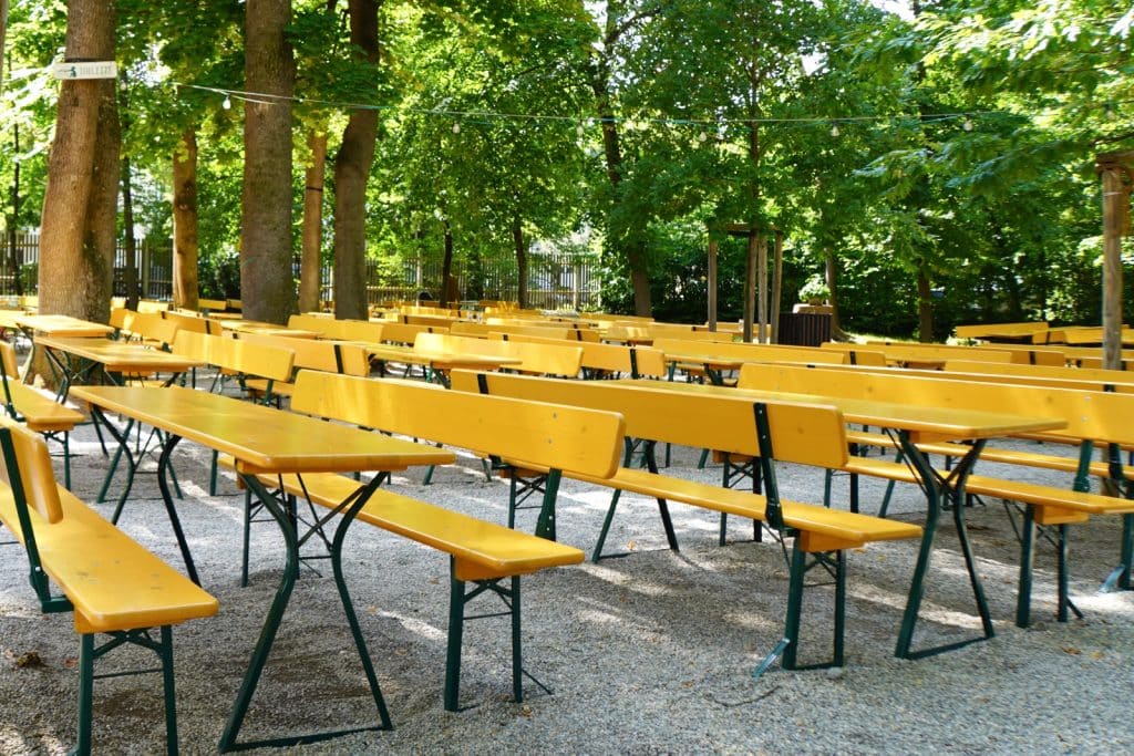 Zahlreiche leere, gelbe Bierbankgarnituren stehen im schattigen Außenbereich des Taxisgartens in München unter hohen, grünen Bäumen auf Kiesboden.